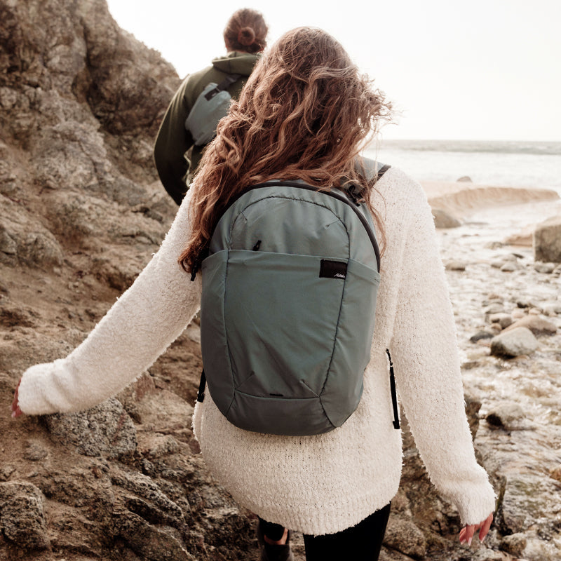 Two women walking along rocky beach. Woman in foreground is wearing a blue packable refraction bag, woman in background is wearing a blue packable refraction sling