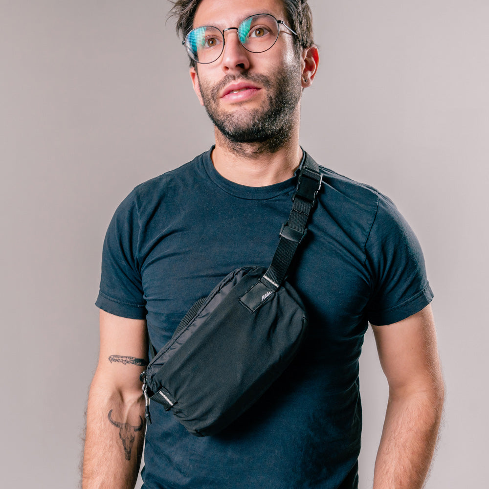 man on light gray background wearing black sling bag across the front of his body