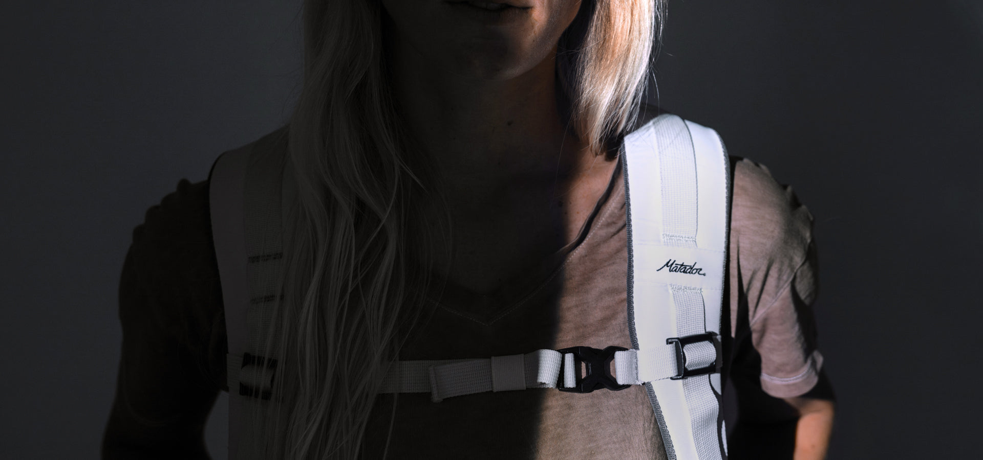 shadowy image of woman wearing white matador backpack with light focusing on the Matador logo shoulder strap