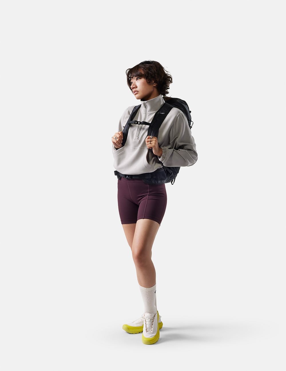 Person wearing a gray jacket, maroon shorts, and yellow shoes with a black backpack on a white background