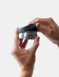 Person holding a metallic container with a black lid on a light gray background