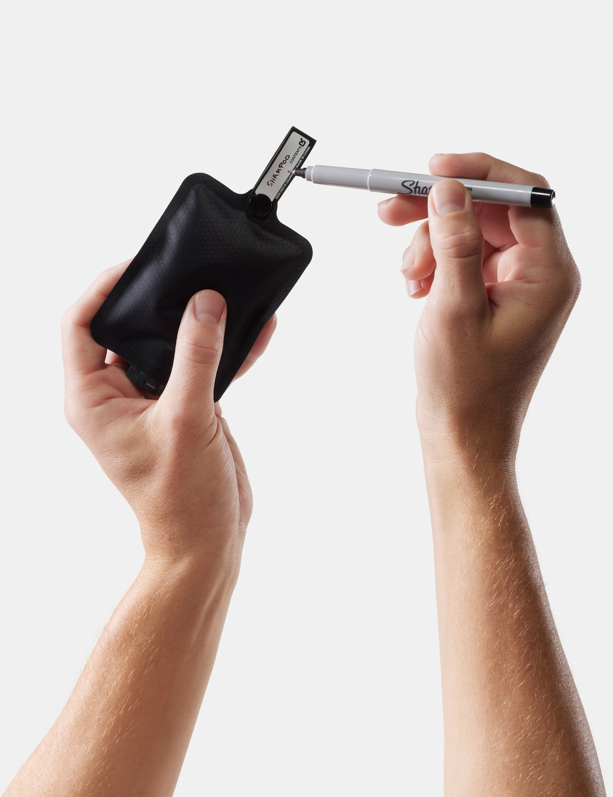 Person using a black Sharpie marker to write "shampoo" on toiletry bottle on a light gray background