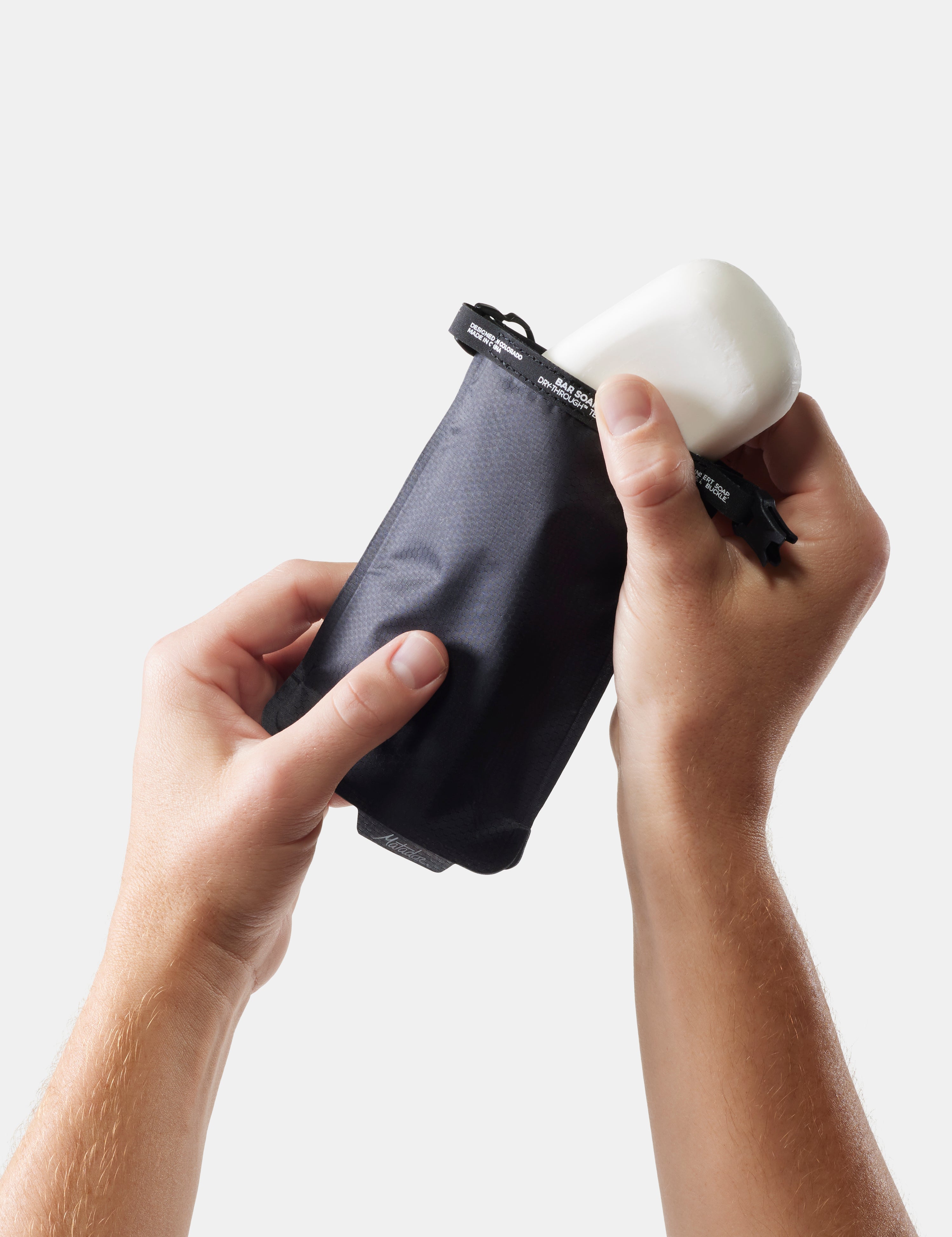 Person holding a black travel pouch and a white soap with a black handle against a light gray background