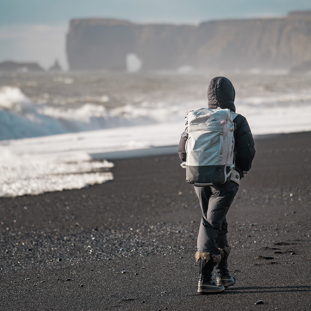 person walking on black sand beach wearing white backpack