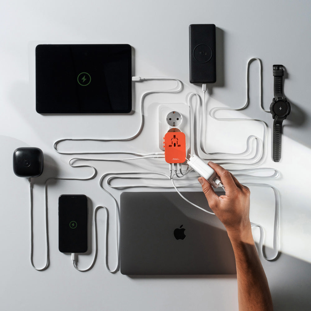 shadowy photo of orange travel adapter plugged into EU outlet with 6 items plugged in: tablet, power bank, smart watch, earbuds, phone, and apple computer.
