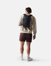 Person wearing a beige sweatshirt and maroon shorts with a black backpack on a white background
