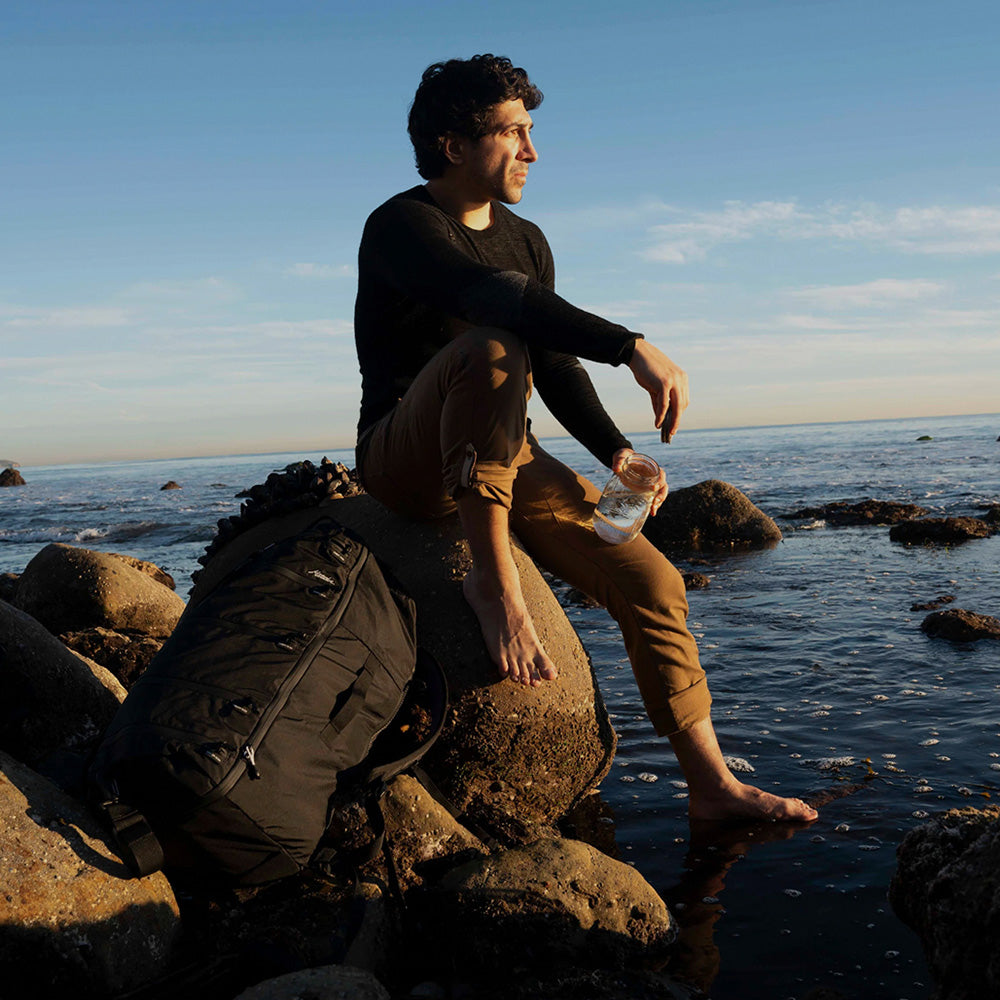 man sitting on rocky shore with black backpack