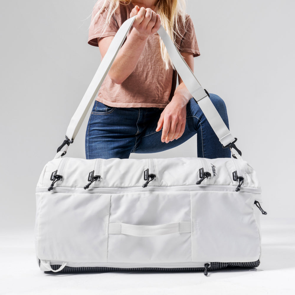 woman kneeling on white backdrop, holding up duffle strap of laid down white seg45 